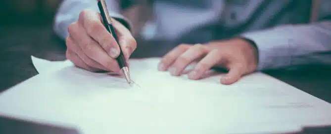 Close-up of a person in a blue shirt writing on documents with a pen, representing the process of applying for business loans.