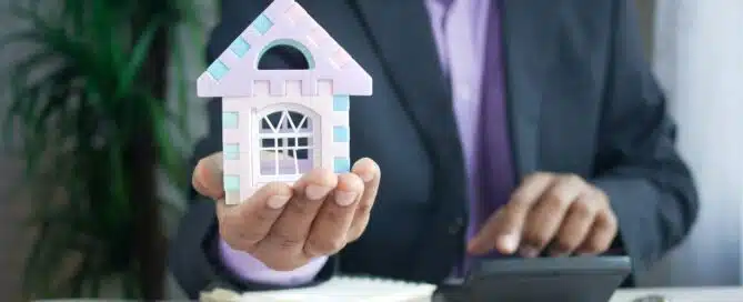 A businessman in a suit holds a small, colorful model house in his hand, representing a commercial mortgage, while using a calculator at a desk with documents and a pen.