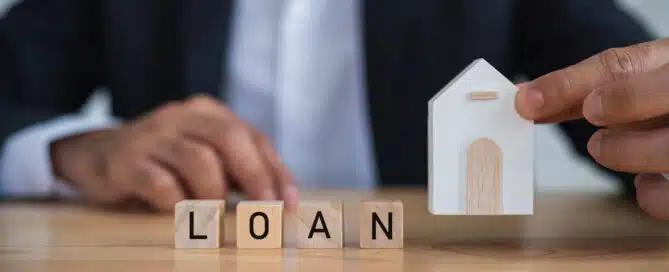 Businessman holding a model house and wooden blocks spelling "LOAN," symbolising real estate or property loans and finance.