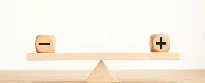 The image shows a wooden balance scale with two blocks on either side. One block has a minus sign (-) and the other has a plus sign (+), representing the concept of weighing pros and cons.